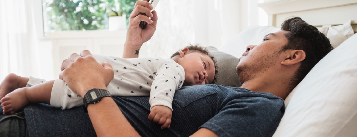 A photographic style image of a man on a bed with his baby on his chest sleeping. The man is viewing his mobile device.