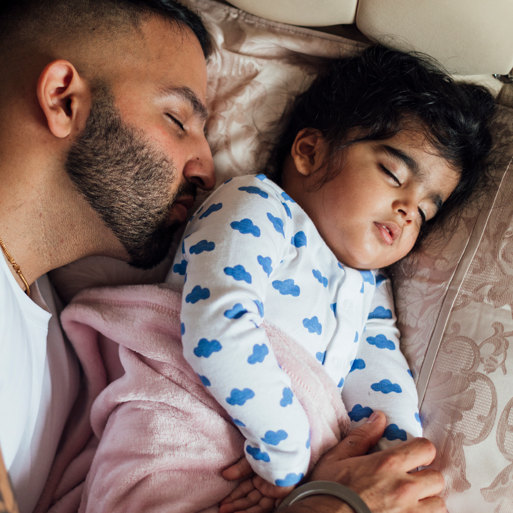 A photographic style image of a father asleep next to his child who is also sleeping.